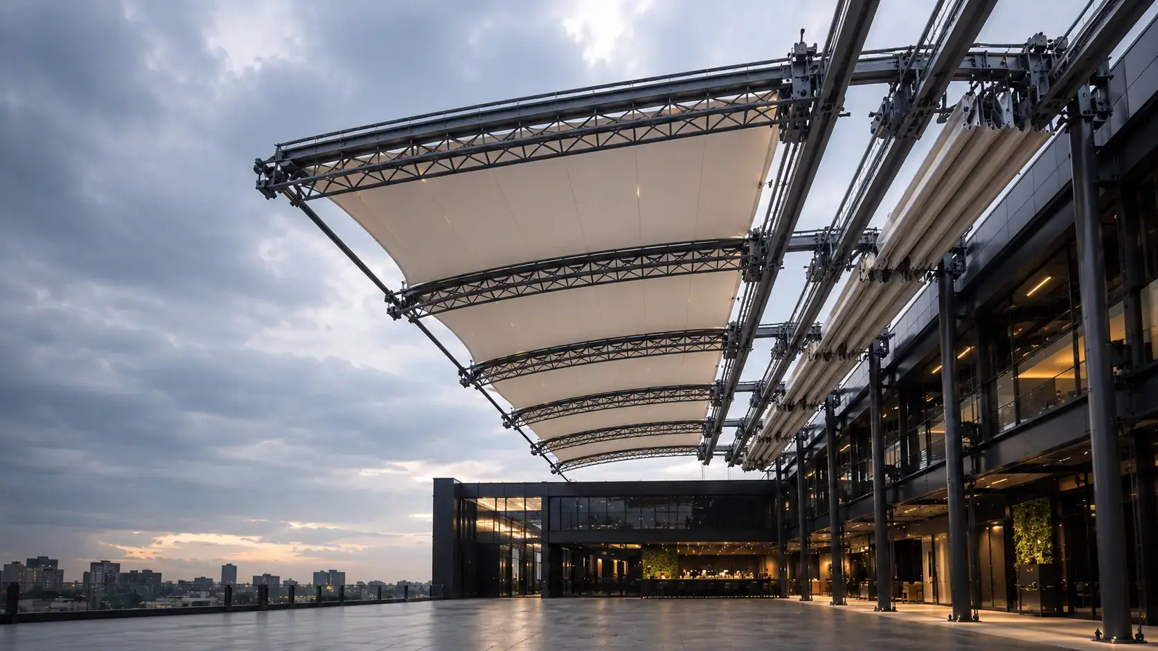 A large-span retractable tensile membrane roof on a truss-and-rail system over a rooftop terrace, membrane bays partly open against a dusk sky